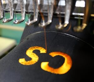 Embroidery machine stitching glowing orange “SC” on black fabric by Righteous Clothing Agency.