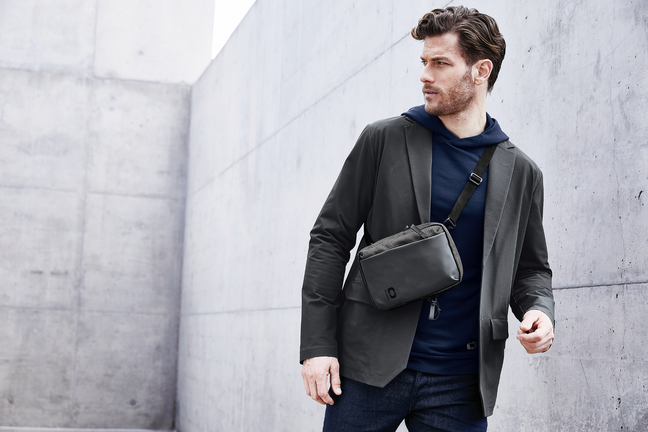 Righteous Clothing Agency model in a charcoal blazer over a navy hoodie with a crossbody bag against a concrete wall.