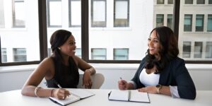 Two women in the Righteous Clothing Agency office meeting at a table, smiling and taking notes.