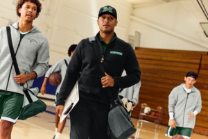 Coach in gym wearing Righteous Clothing Agency black quarter-zip and cap, carrying clipboard and duffel.