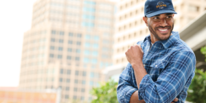 Smiling man in blue plaid shirt and logo cap outdoors, styled by Righteous Clothing Agency.