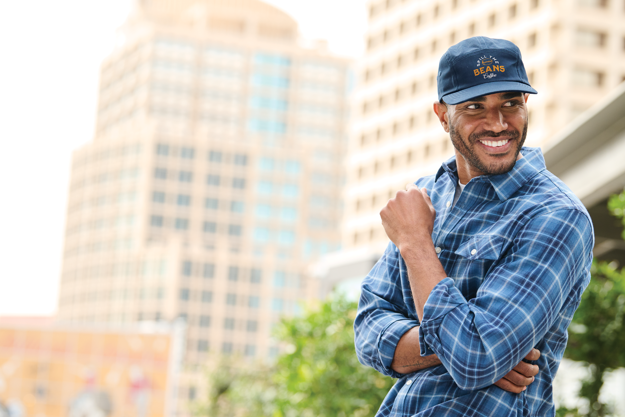Smiling man in blue plaid shirt and logo cap outdoors, styled by Righteous Clothing Agency.