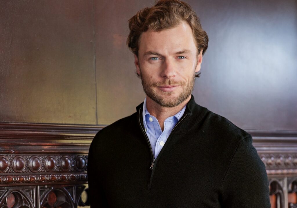 Man in a black zip-up sweater over a blue shirt, standing in front of a wooden background, wearing Righteous apparel.
