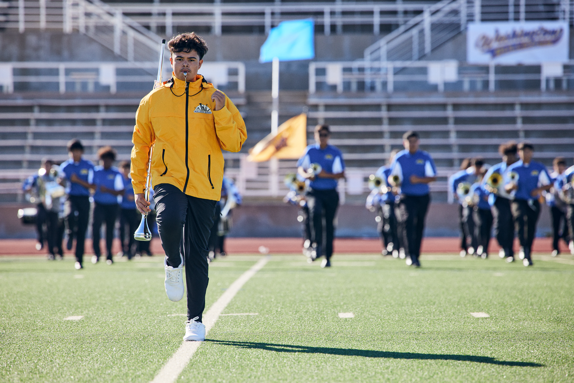 Righteous branded yellow jacket worn by a coach leading a marching band on a football field.