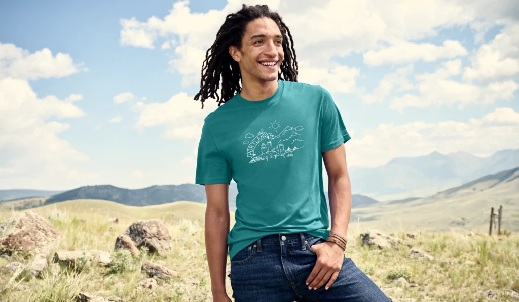 Man in a Righteous teal graphic tee stands in a sunny mountain field.