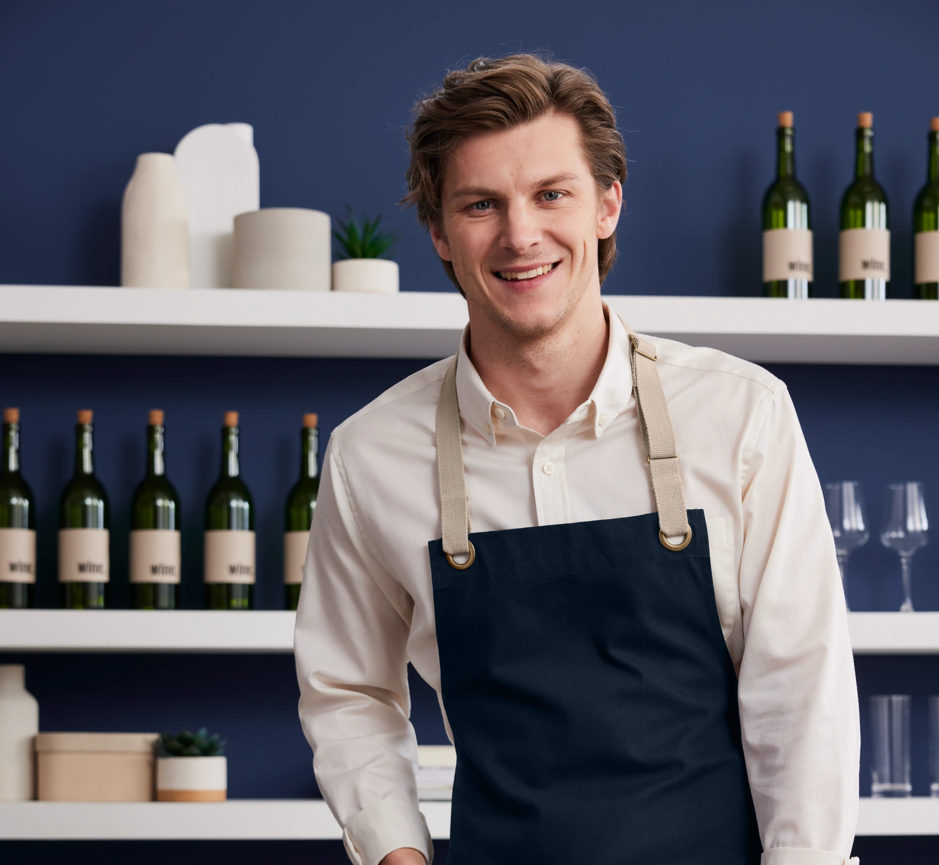Server in a white shirt and navy apron from Righteous Clothing Agency stands by wine bottles. 