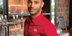 Bartender in a red embroidered uniform from Righteous Clothing Agency stands beside beer taps.