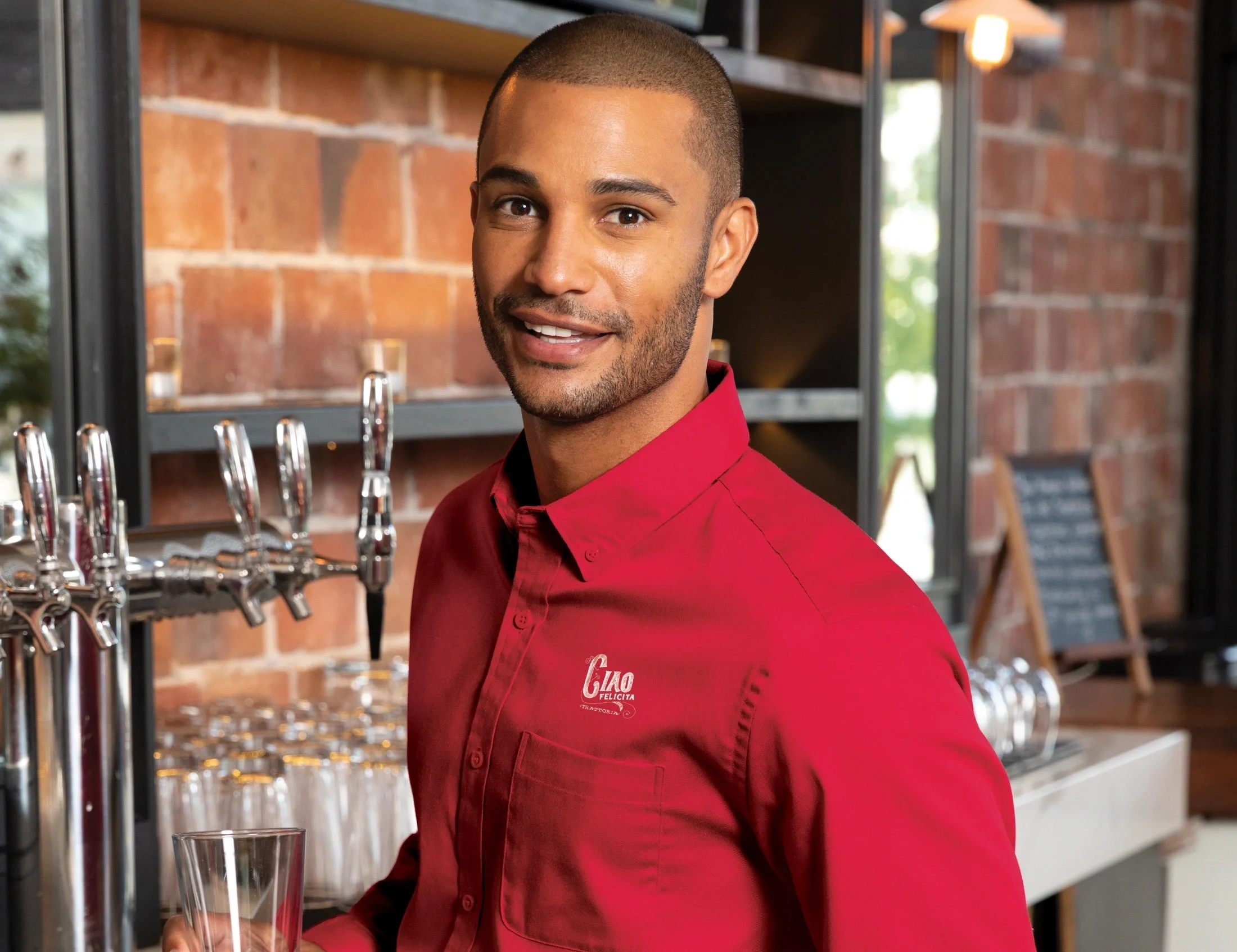 Bartender in a red embroidered uniform from Righteous Clothing Agency stands beside beer taps.